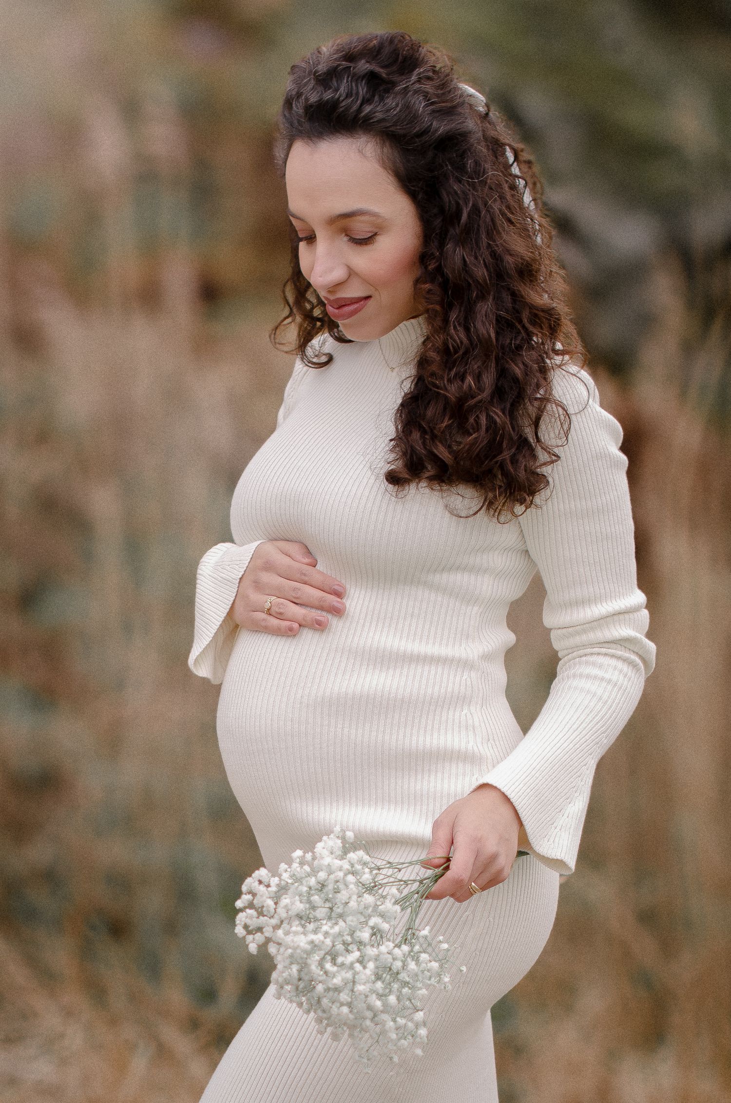 Pregnant woman in white ribbed dress holding baby's breath flowers while posing outdoors in nature.