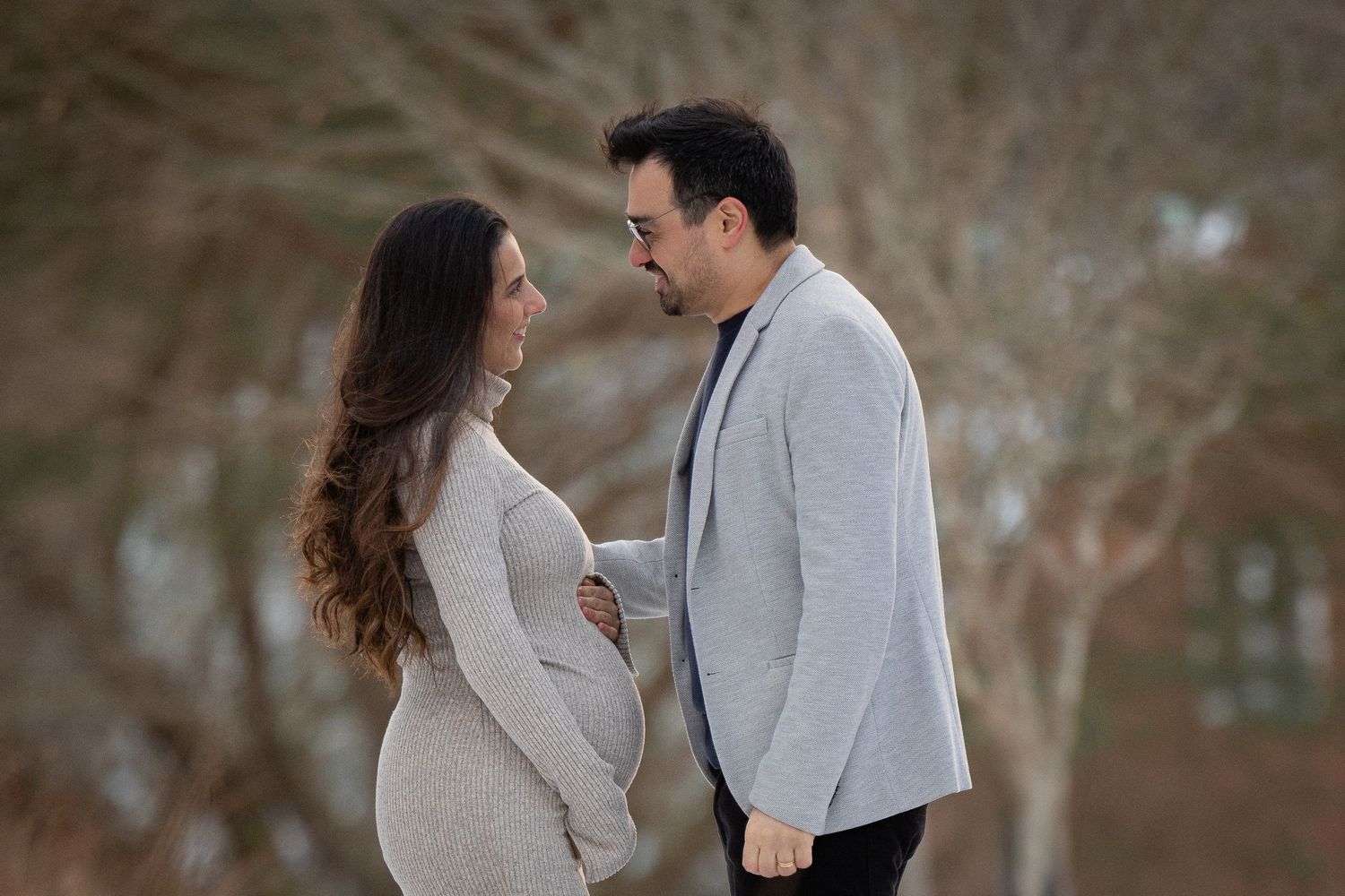 Expecting couple facing each other tenderly outdoors in winter, man in grey blazer with pregnant woman in knit dress.