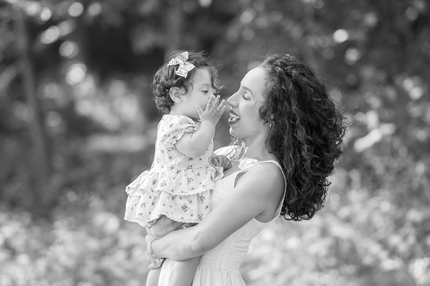 Black and white photo of a curly-haired mother lifting and laughing with her baby girl outdoors.
