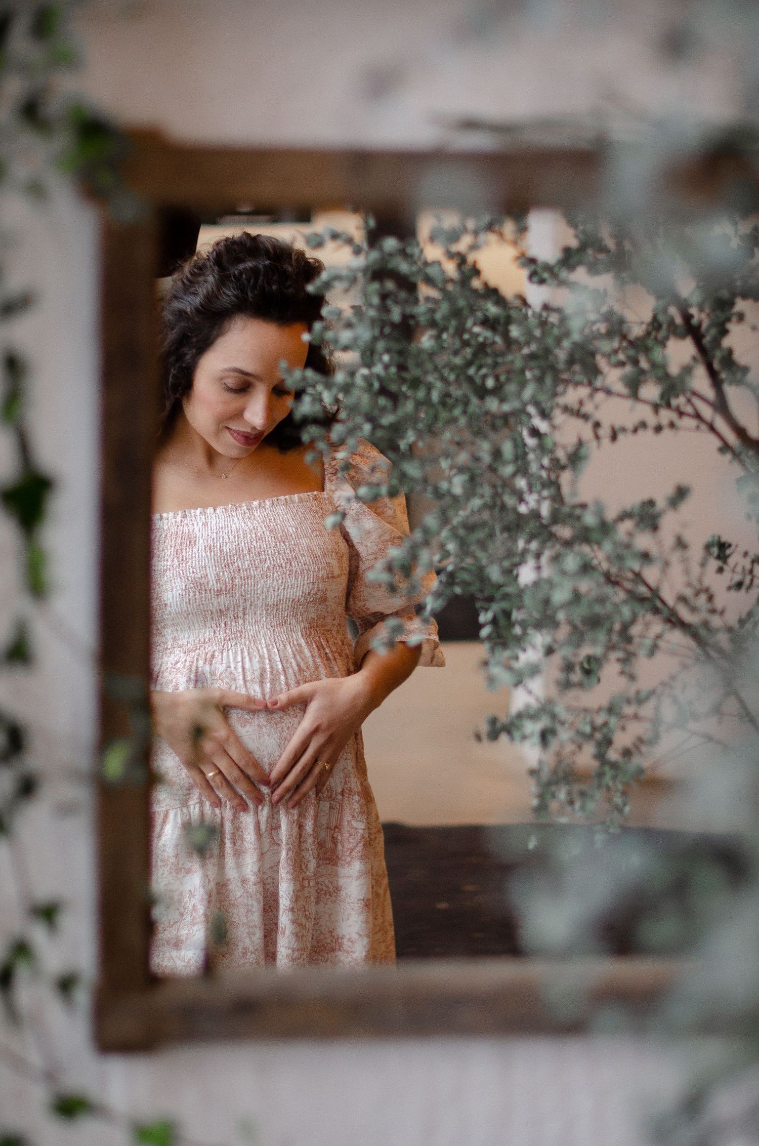 Pregnant woman in floral dress cradling baby bump framed by eucalyptus branches indoors.