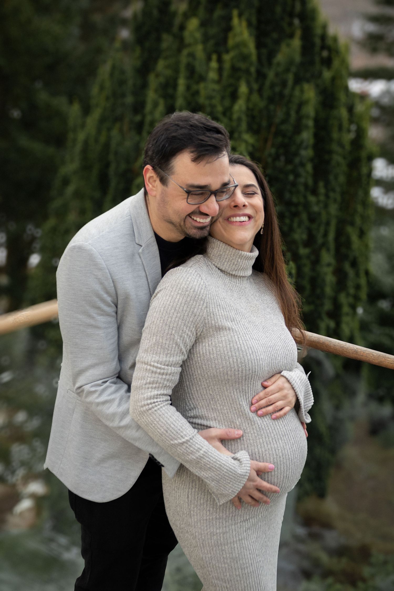 Smiling couple outdoors, man embracing pregnant woman in grey turtleneck dress near evergreen trees.