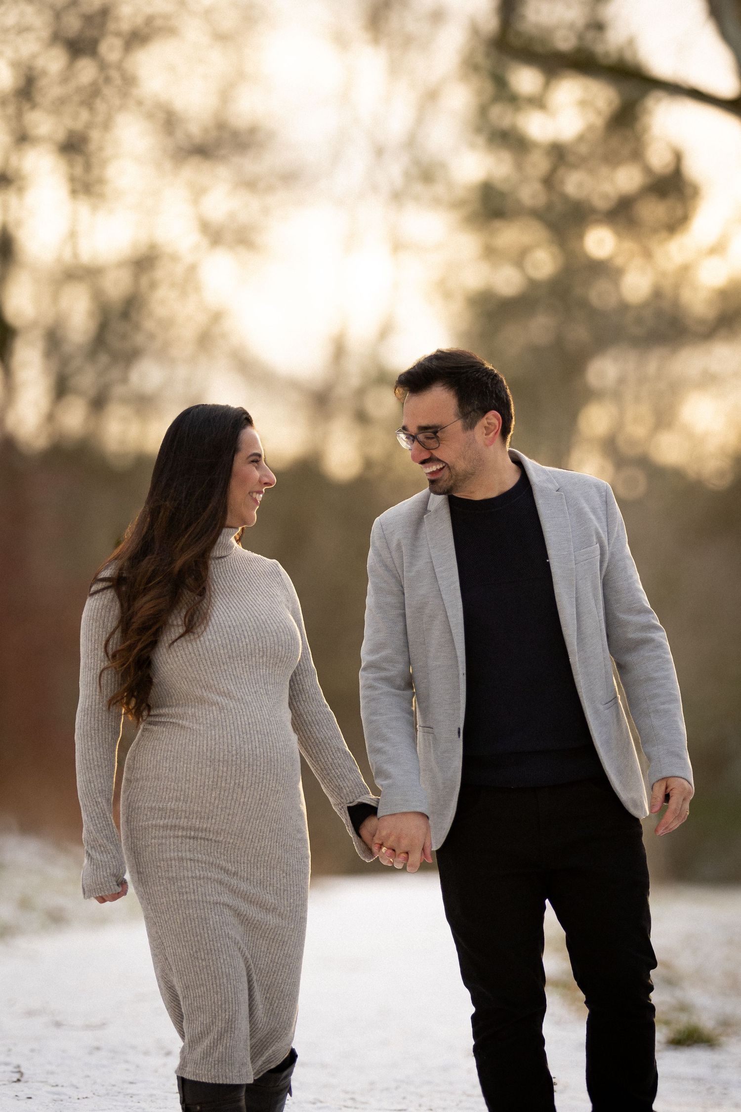 Happy pregnant couple holding hands and smiling at each other while walking in snowy winter park.