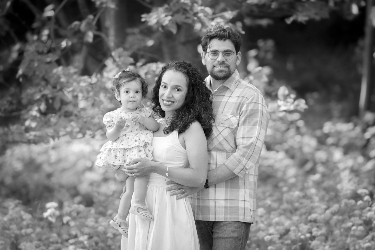 Black and white family portrait of smiling couple holding toddler daughter outdoors in garden setting.