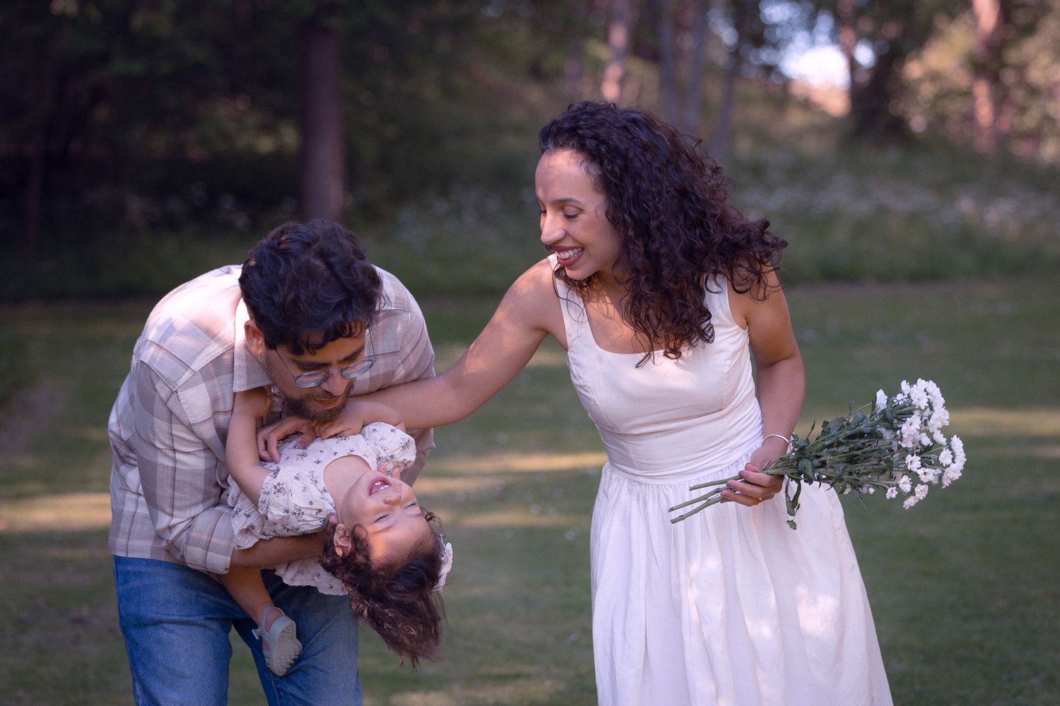 Playful family moment as father dips laughing toddler while mother in white dress holds white flowers in park.