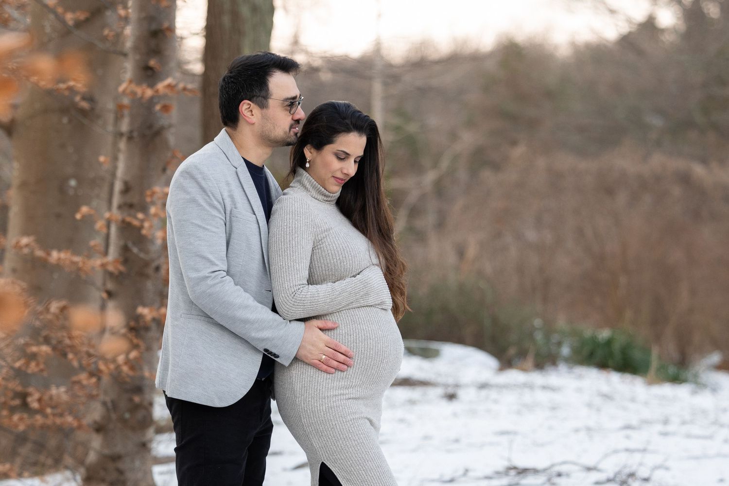 Couple embracing in snowy winter park, man holding pregnant woman's belly among autumn trees.