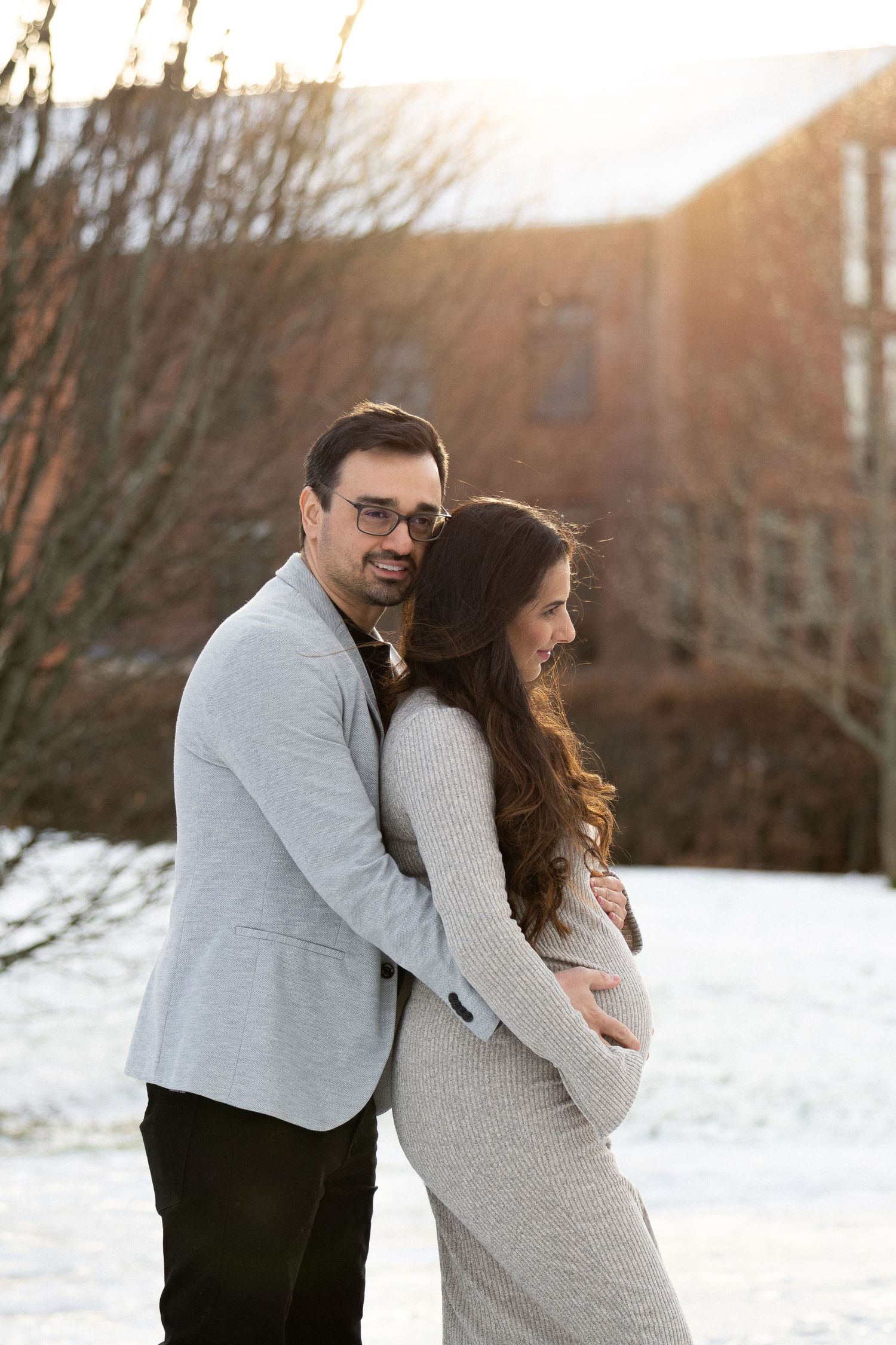 Couple embracing in snowy winter setting with golden sunlight, man hugging pregnant woman from behind outdoors.
