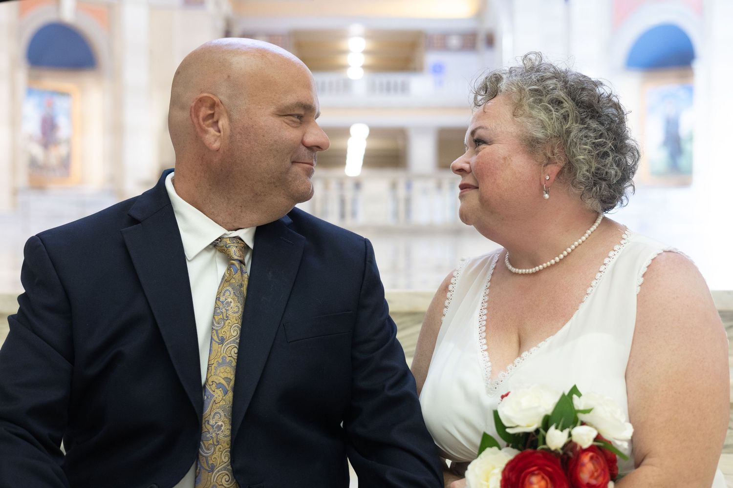 Couple in wedding attire smiling at each other, groom in navy suit, bride in white dress holding red bouquet.