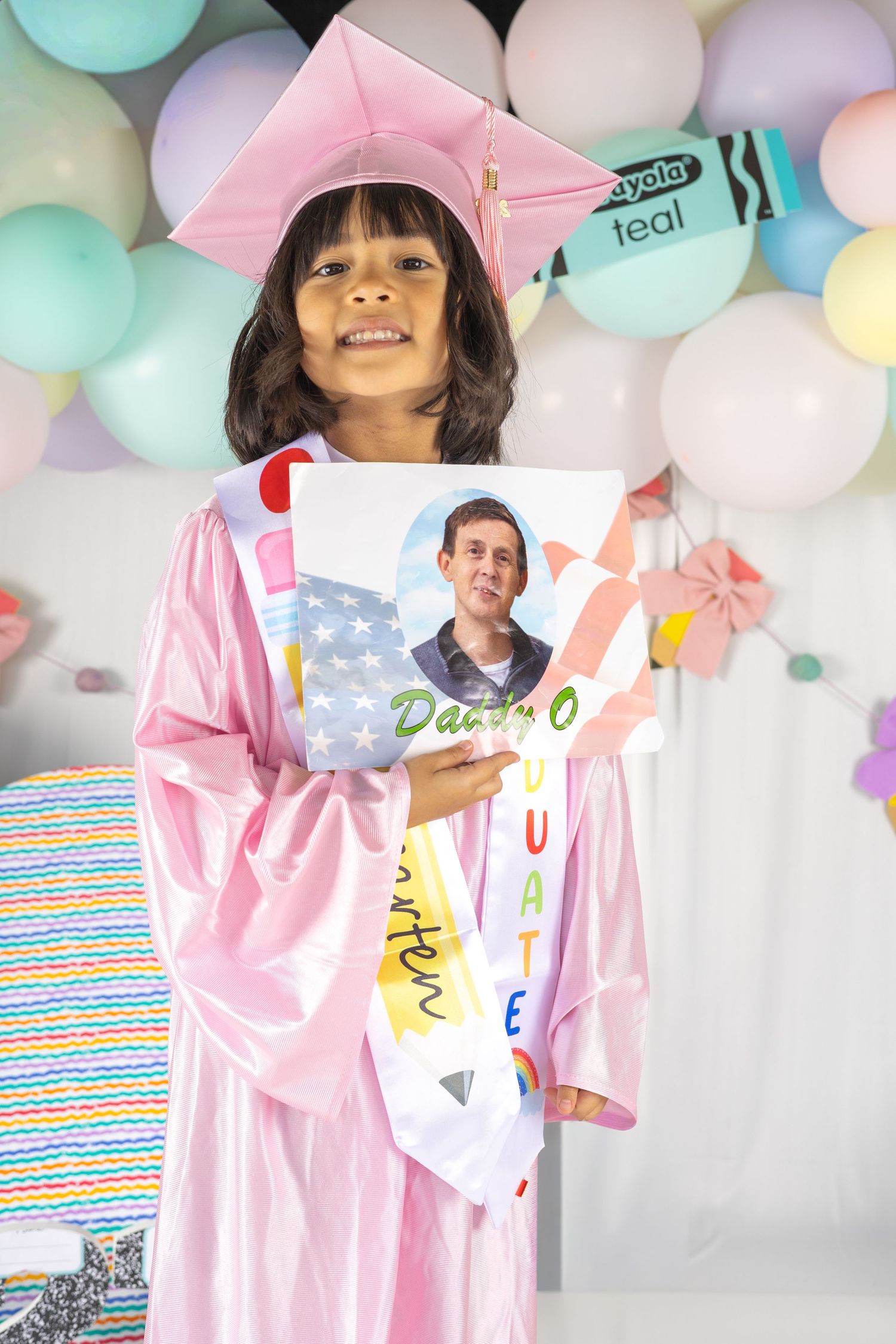 Young girl in pink graduation cap and gown holding a graduate photo board, standing in front of colorful balloons.