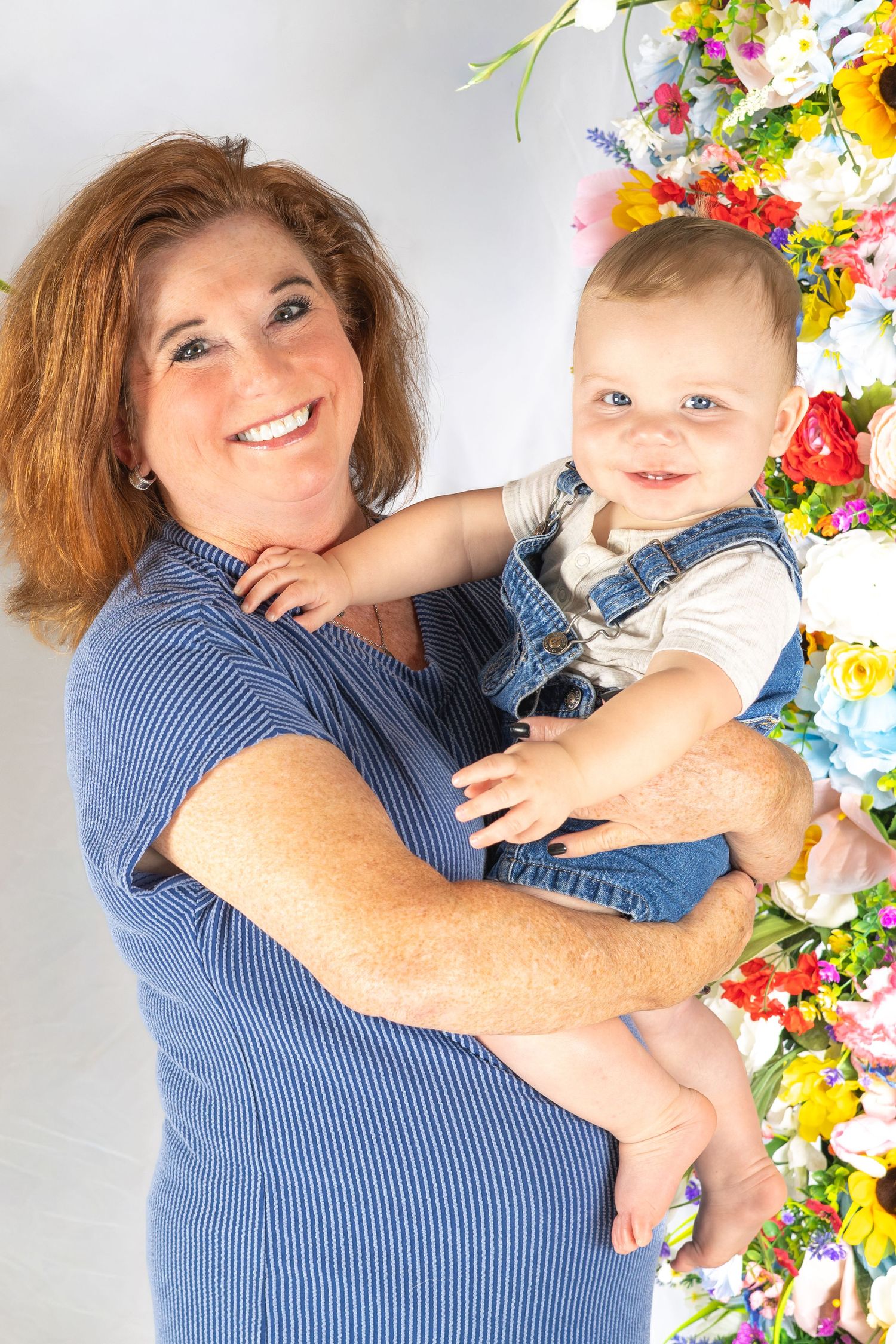 Smiling redhead woman holds happy baby in denim overalls beside vibrant flower wall display.