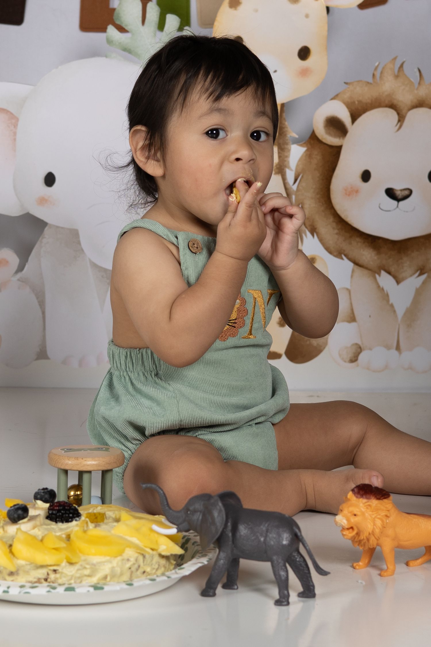 Baby in green romper eating cake at safari-themed first birthday party with toy animals and fruit cake.