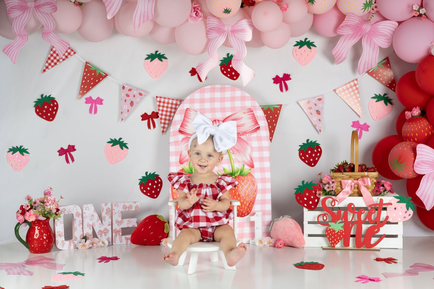 Baby girl sits in pink chair at strawberry-themed first birthday cake smash with balloons and decorations.