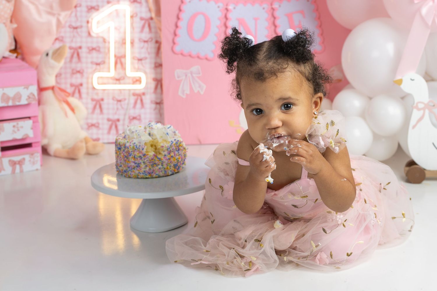 Baby girl in pink tutu dress eating first birthday cake surrounded by pink balloons and decorations.