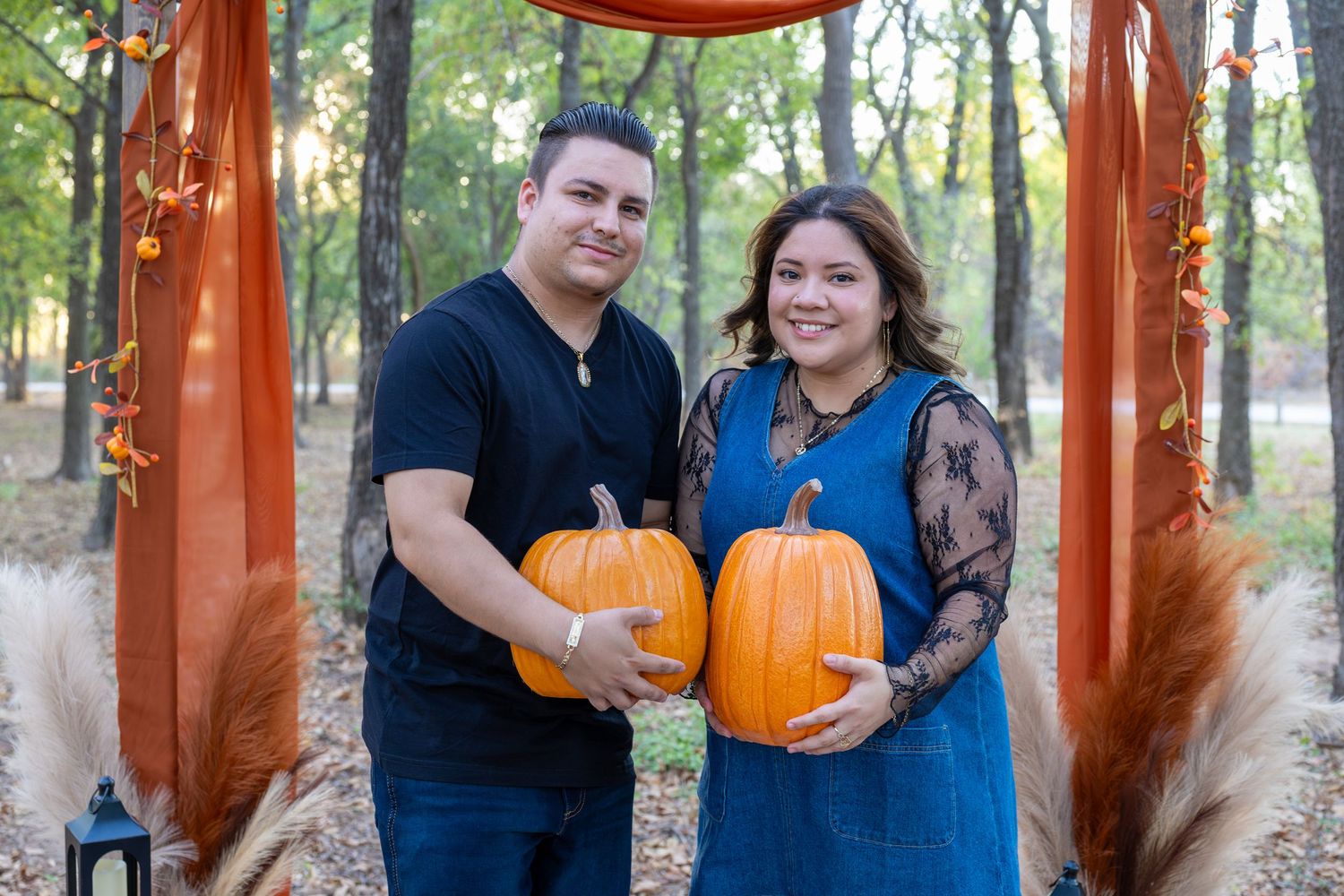 Couple holding orange pumpkins under a rustic fall arch with orange draping in an autumn forest setting.