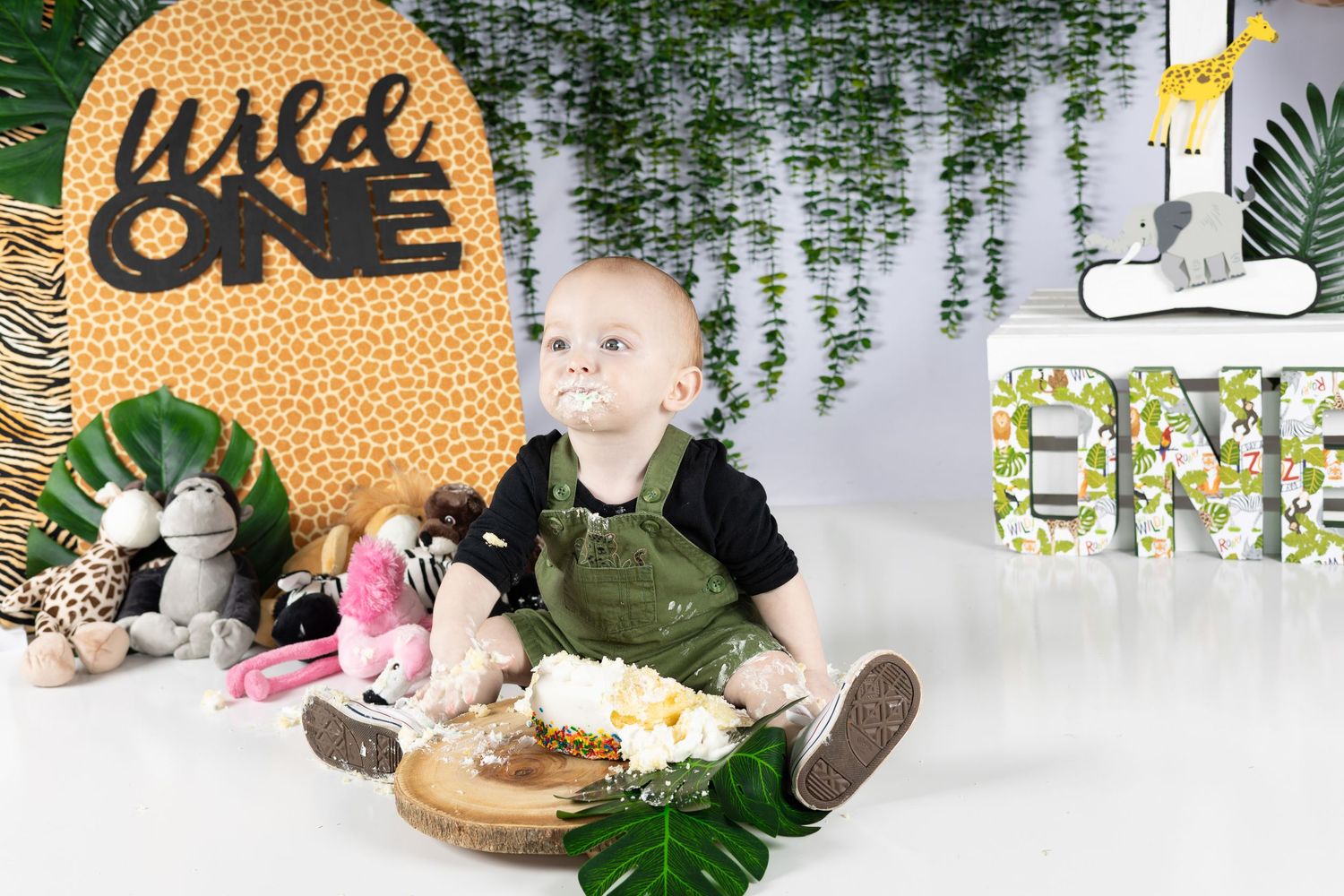 Baby boy in overalls sits with smash cake at Wild One jungle safari first birthday party photo session.