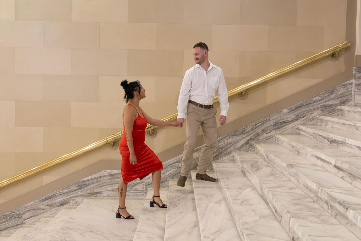 A man and woman dance together on marble stairs, she wearing a red dress and he in white shirt and khaki pants.