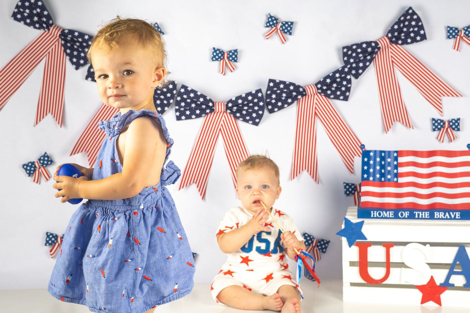 Two toddlers pose before patriotic USA decorations with American flags, bows, and red, white, and blue banners.