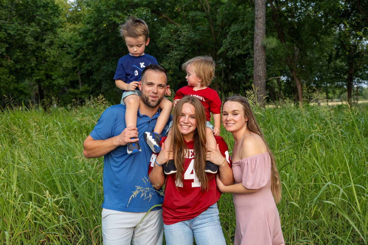 Happy family of five posing outdoors in a lush green field, with two young boys on dad's shoulders.