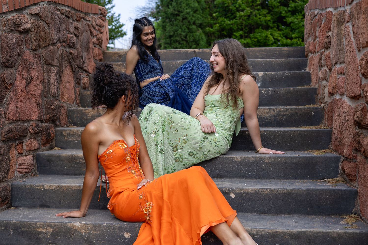 Three women in elegant dresses pose on stone steps outdoors, wearing orange, green, and blue formal gowns.