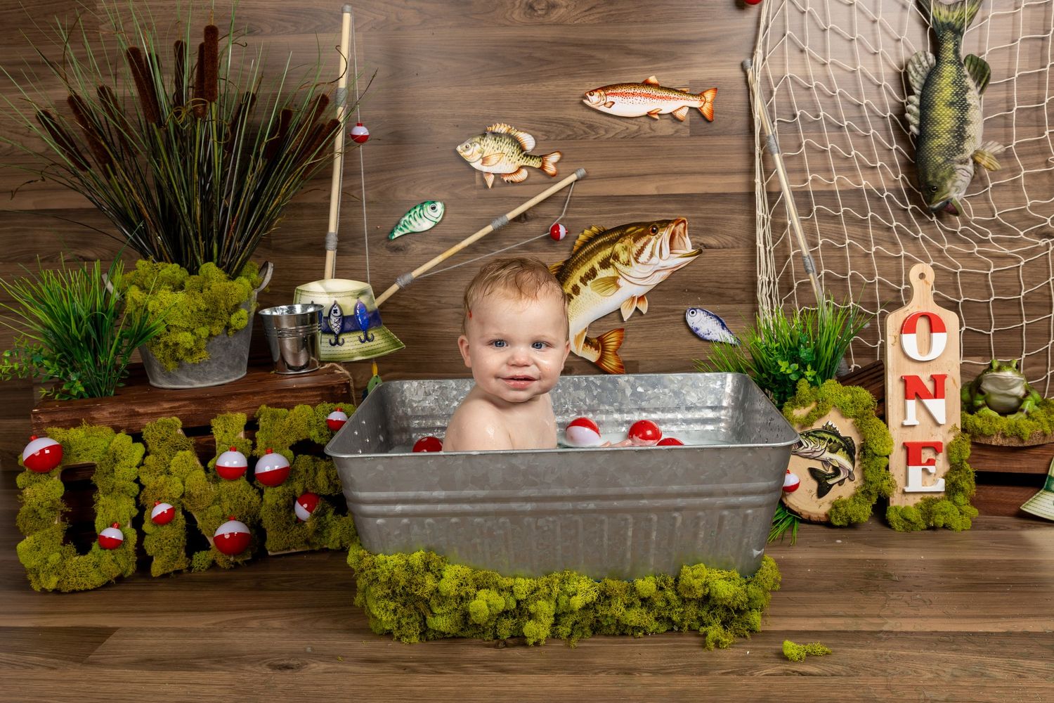 Baby smiling in galvanized tub with fishing-themed first birthday decorations including fish, nets, and ONE sign.