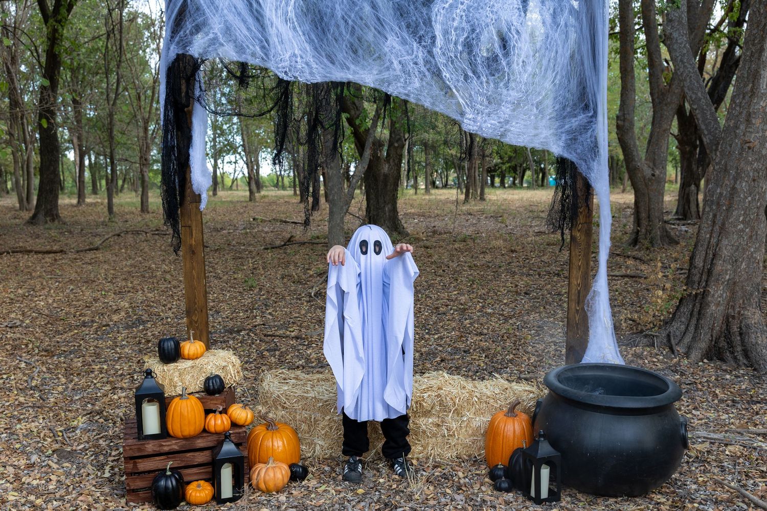 Child in ghost costume stands in Halloween display with pumpkins, hay bales, cauldron, and spooky decorations in a forest.