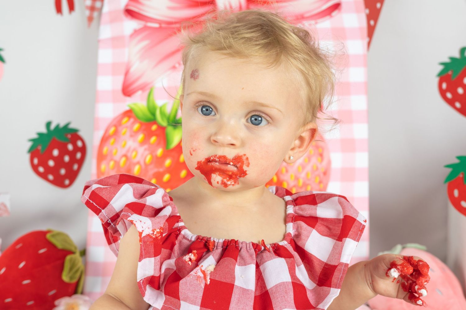 Toddler girl in red gingham dress with strawberry cake smash on her face, celebrating first birthday.