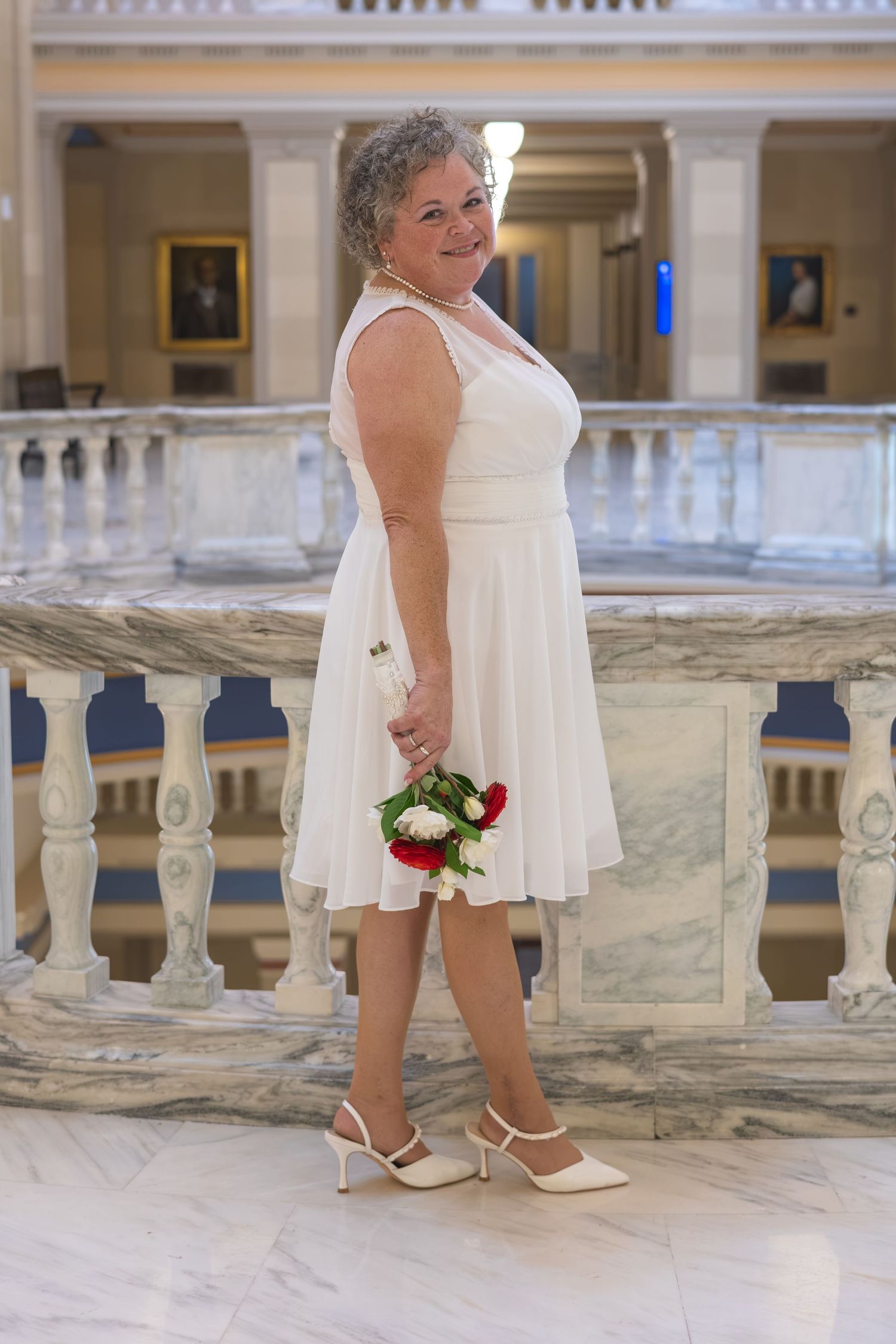 Older bride in white dress holding red and white bouquet poses by marble balustrade in elegant hall.