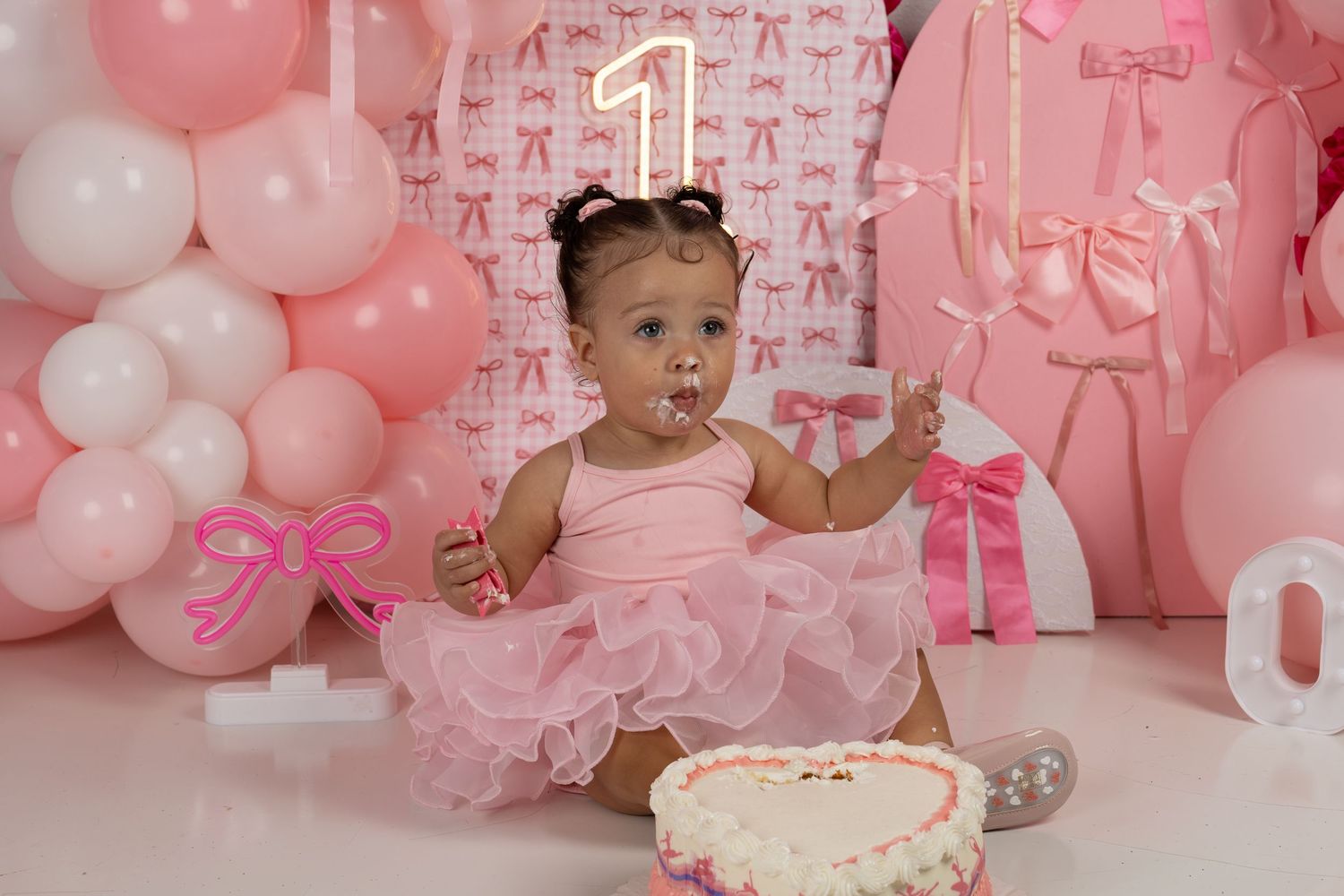 Baby girl in pink tutu celebrates first birthday with cake smash, surrounded by pink balloons and bow decorations.
