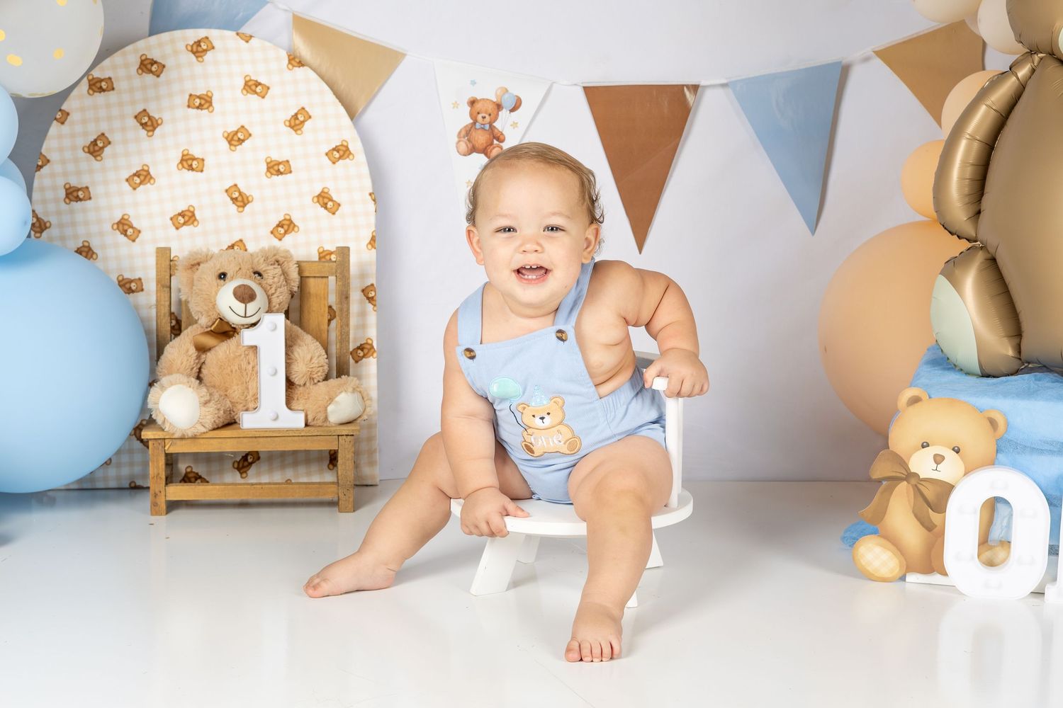 Smiling baby boy in blue romper sits at first birthday teddy bear themed photo shoot with balloons and stuffed animals.