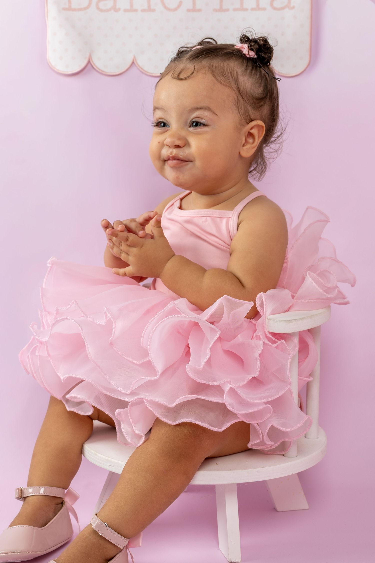 Baby girl in pink ruffle tutu dress sitting on white chair, clapping hands, ballerina birthday party theme.