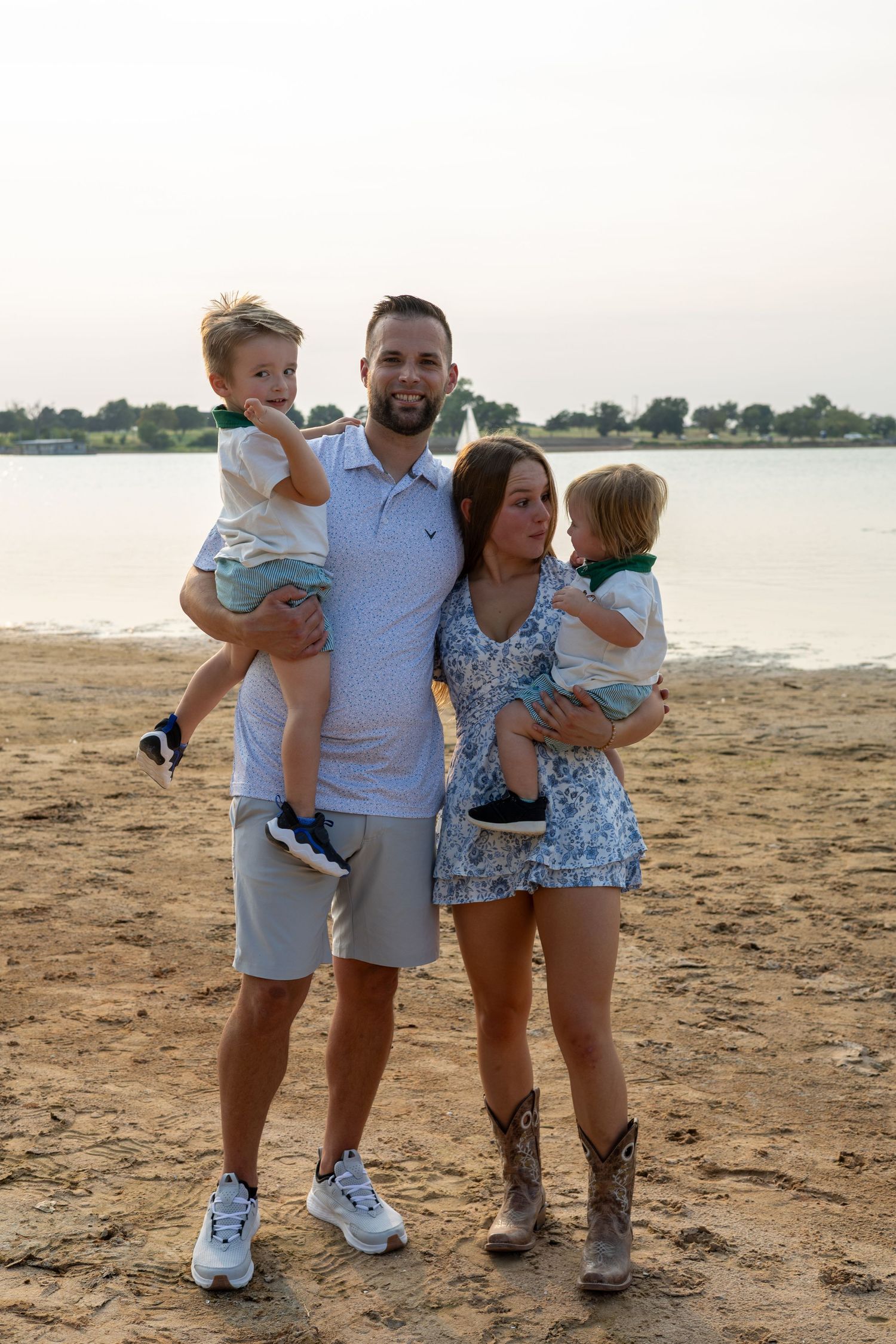 A young family of four poses on a sandy beach at sunset, parents holding two toddlers while smiling for a portrait.