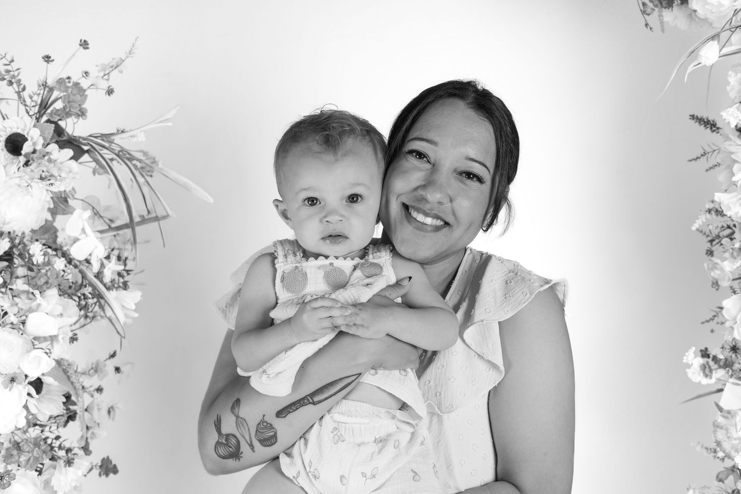 Black and white photo of a smiling woman holding a happy baby girl, surrounded by floral decorations.