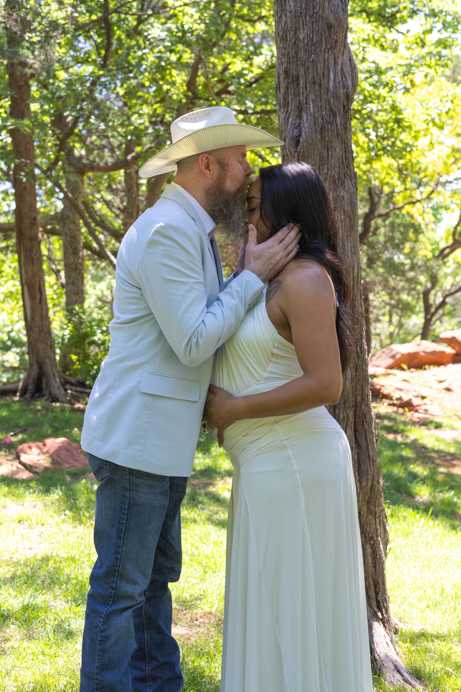 Couple sharing a romantic kiss by a tree in a lush garden, man in cowboy hat and white blazer, woman in cream dress.