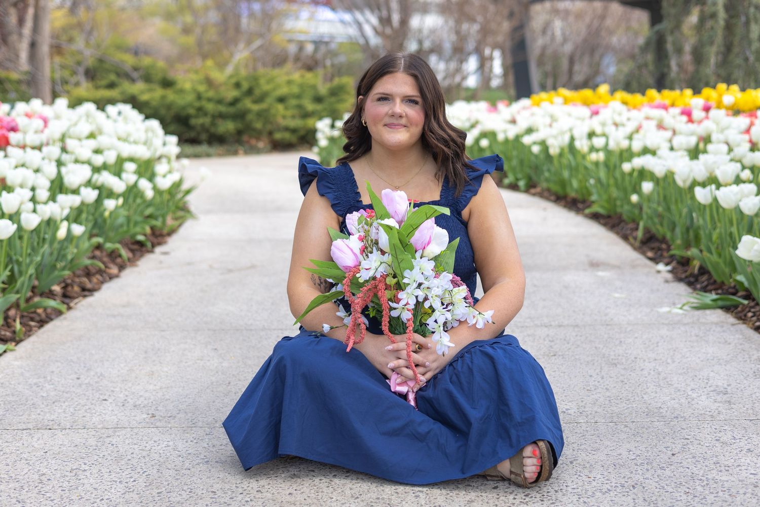 A person in a blue dress sits on a garden path surrounded by blooming white flowers and spring foliage.