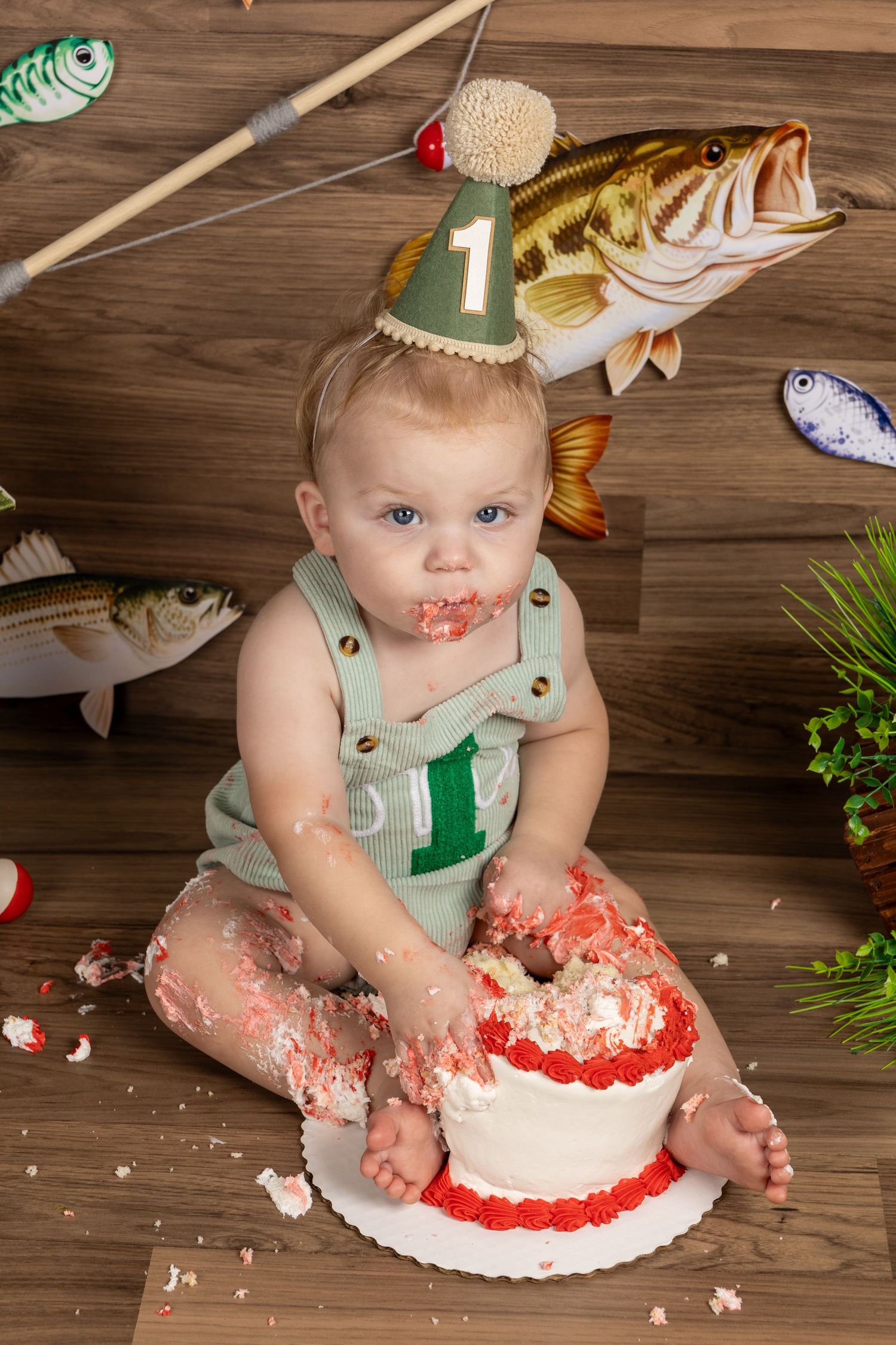 Baby boy in fishing-themed first birthday party smashing cake, wearing number 1 hat, surrounded by fish decorations.
