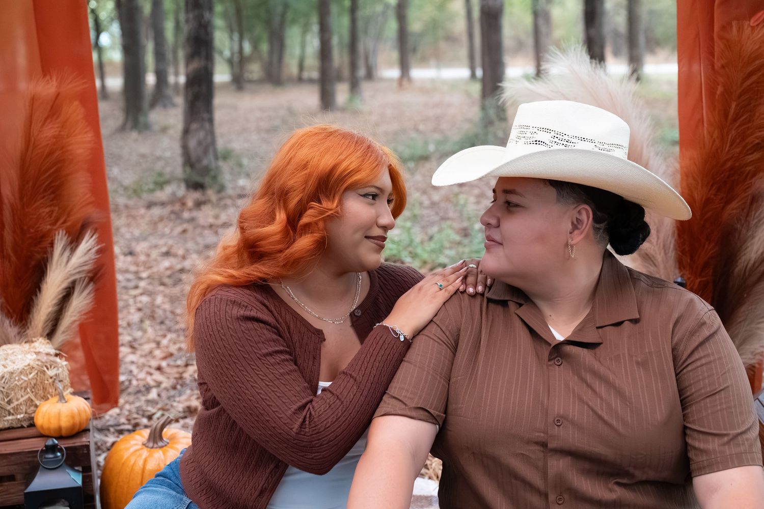 Two women in brown tops share a warm moment outdoors, one with red hair, one in a cowboy hat, near fall pumpkins.