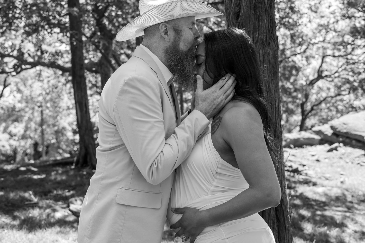 Black and white photo of a couple sharing a romantic kiss outdoors near a tree, man wearing a cowboy hat and suit.