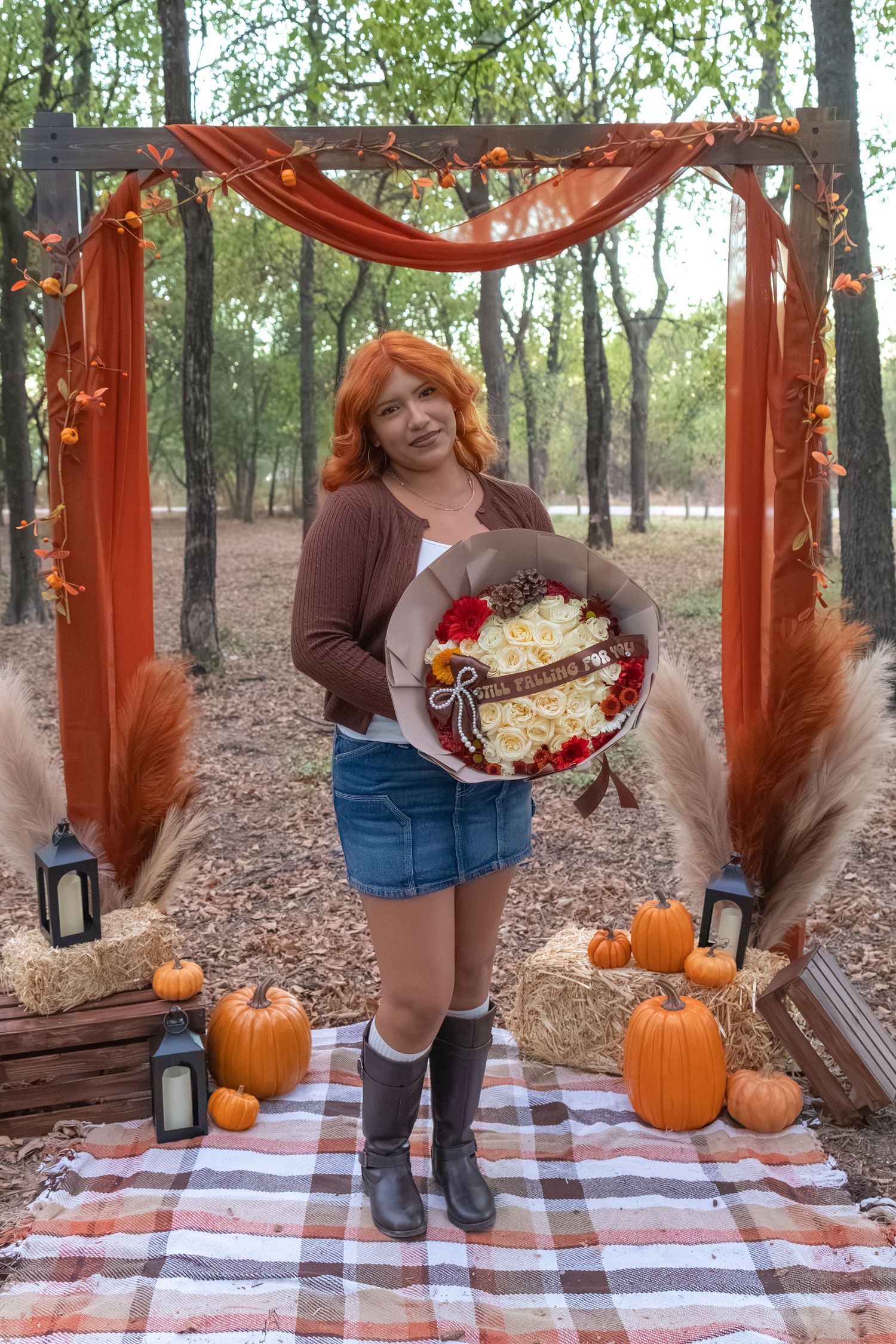 Woman with red hair holds floral bouquet under rustic autumn arch with pumpkins and lanterns in forest setting.