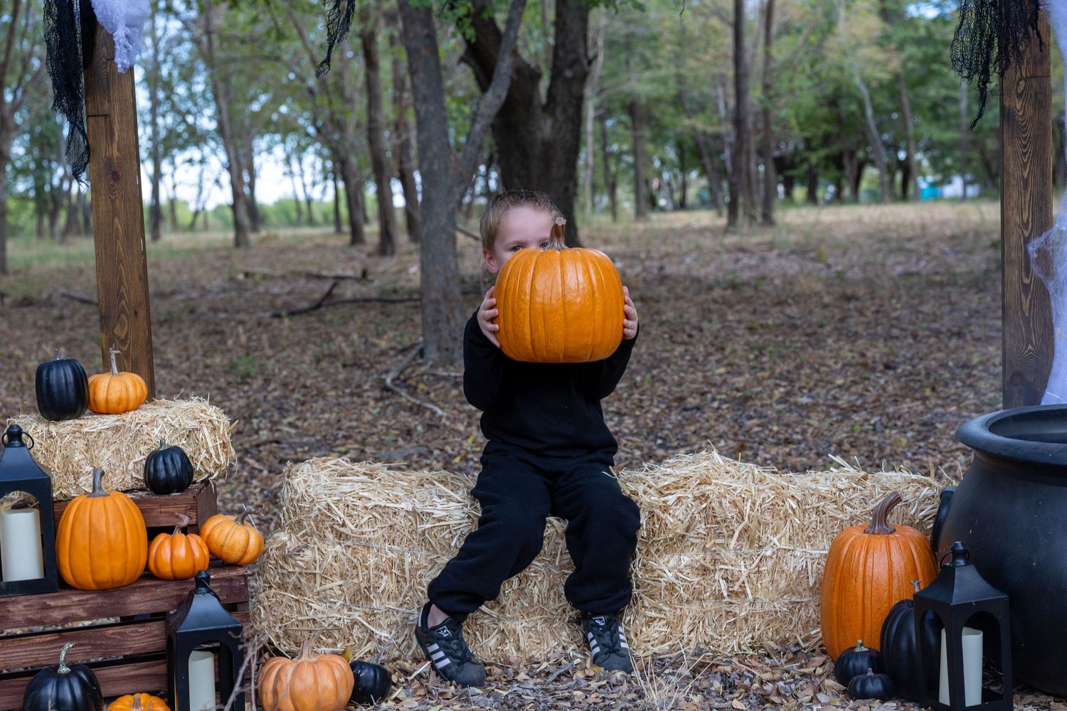 Child in black outfit lifting large orange pumpkin surrounded by hay bales and pumpkins in an outdoor fall Halloween setting.