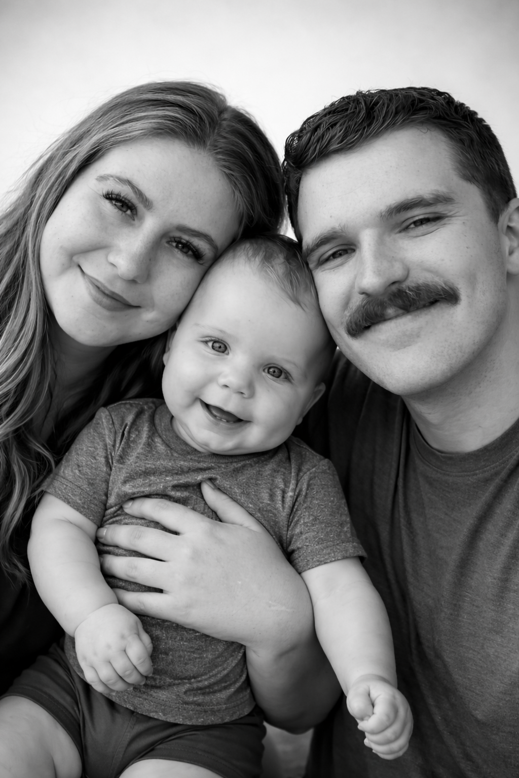 Black and white family portrait of smiling parents with their happy laughing baby in a gray onesie.
