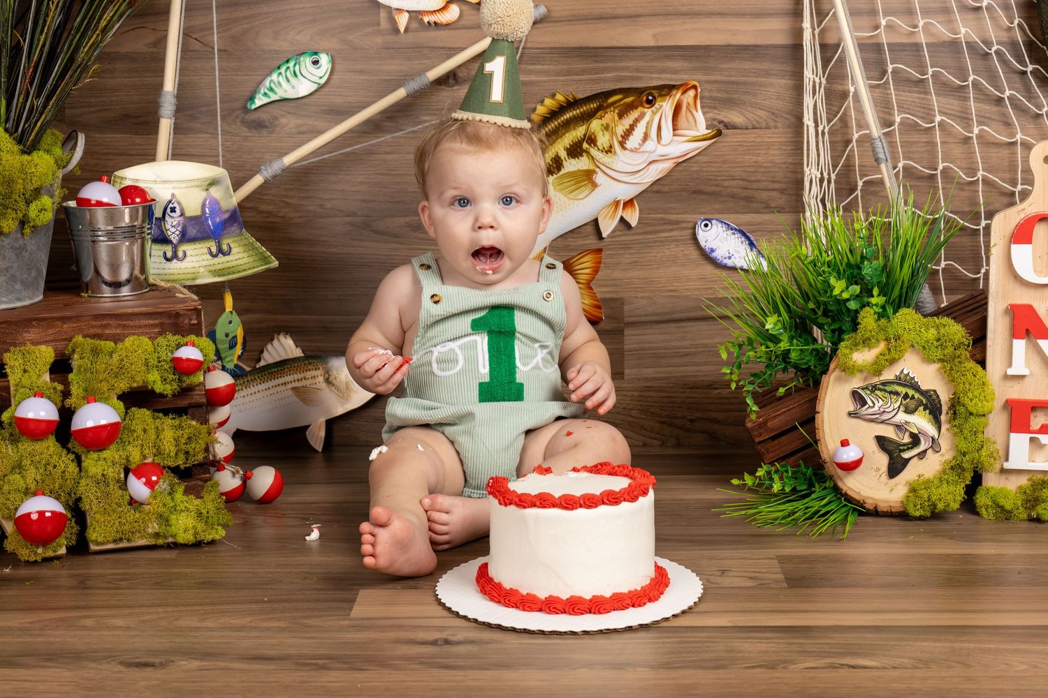 Baby's first birthday cake smash photo with fishing theme decorations, fish, bobbers, and net backdrop.