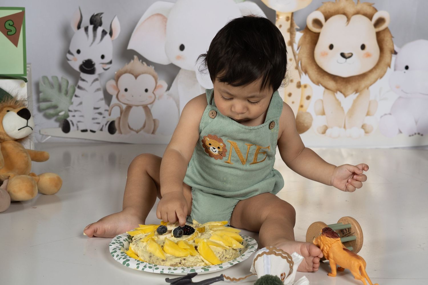 Baby in safari romper sits with fruit plate surrounded by jungle animal toys and decorations at first birthday party.
