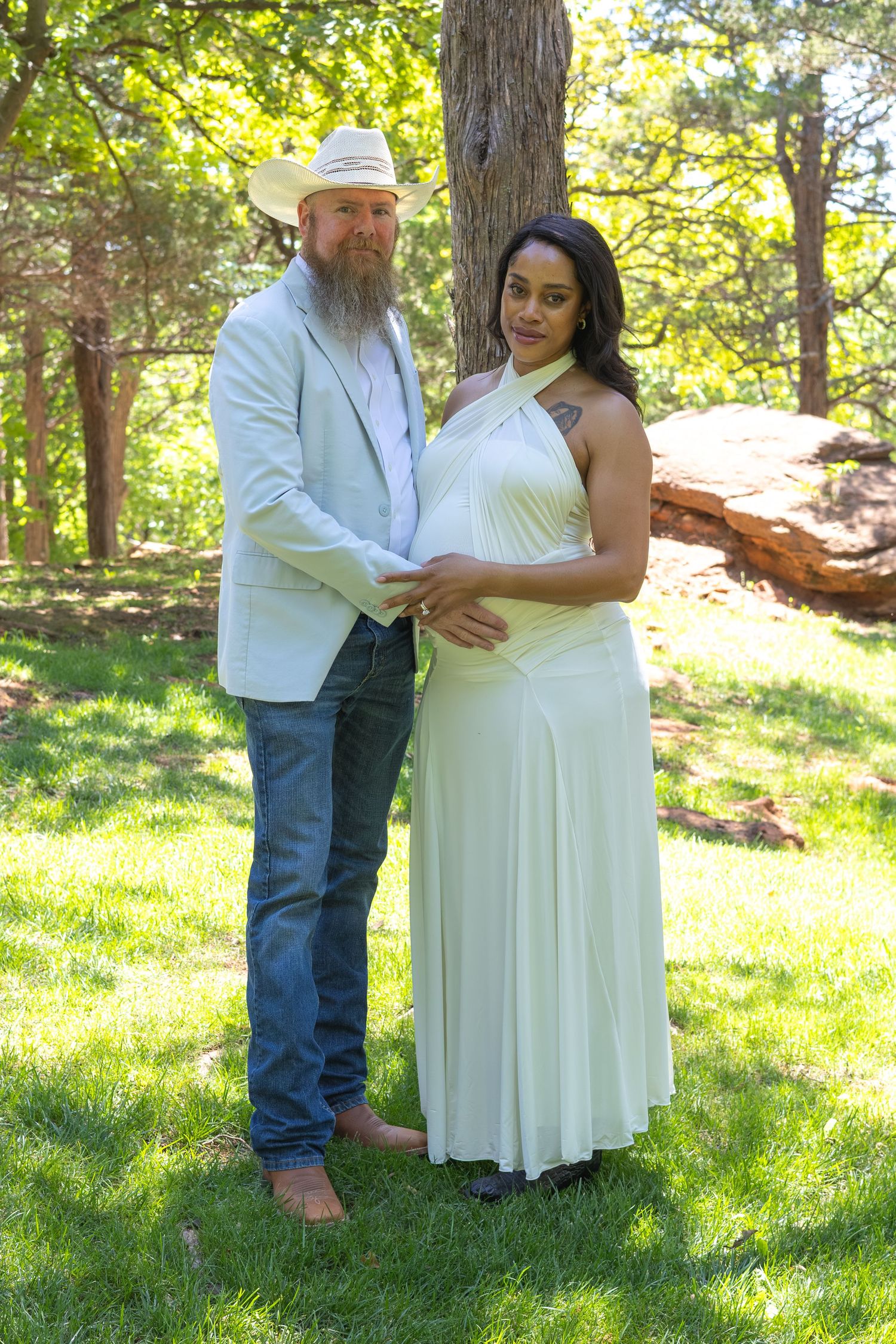 Pregnant couple posing outdoors in green park, man in white blazer and cowboy hat, woman in elegant white halter dress.