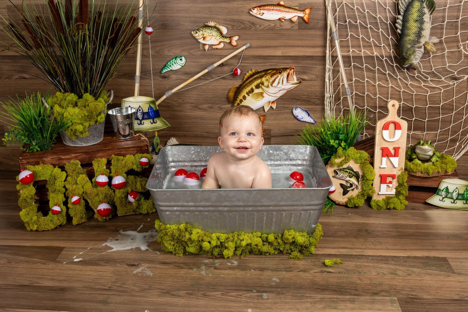 Baby smiling in a tin tub with fishing-themed first birthday decor, moss letters, fish props, and a wooden backdrop.