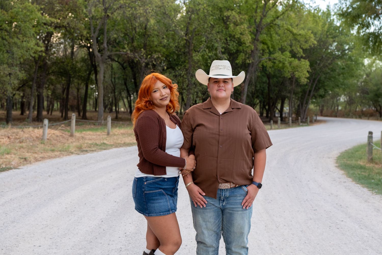 A couple poses on a rural gravel road, woman with red hair in denim skirt, man in cowboy hat and brown shirt.