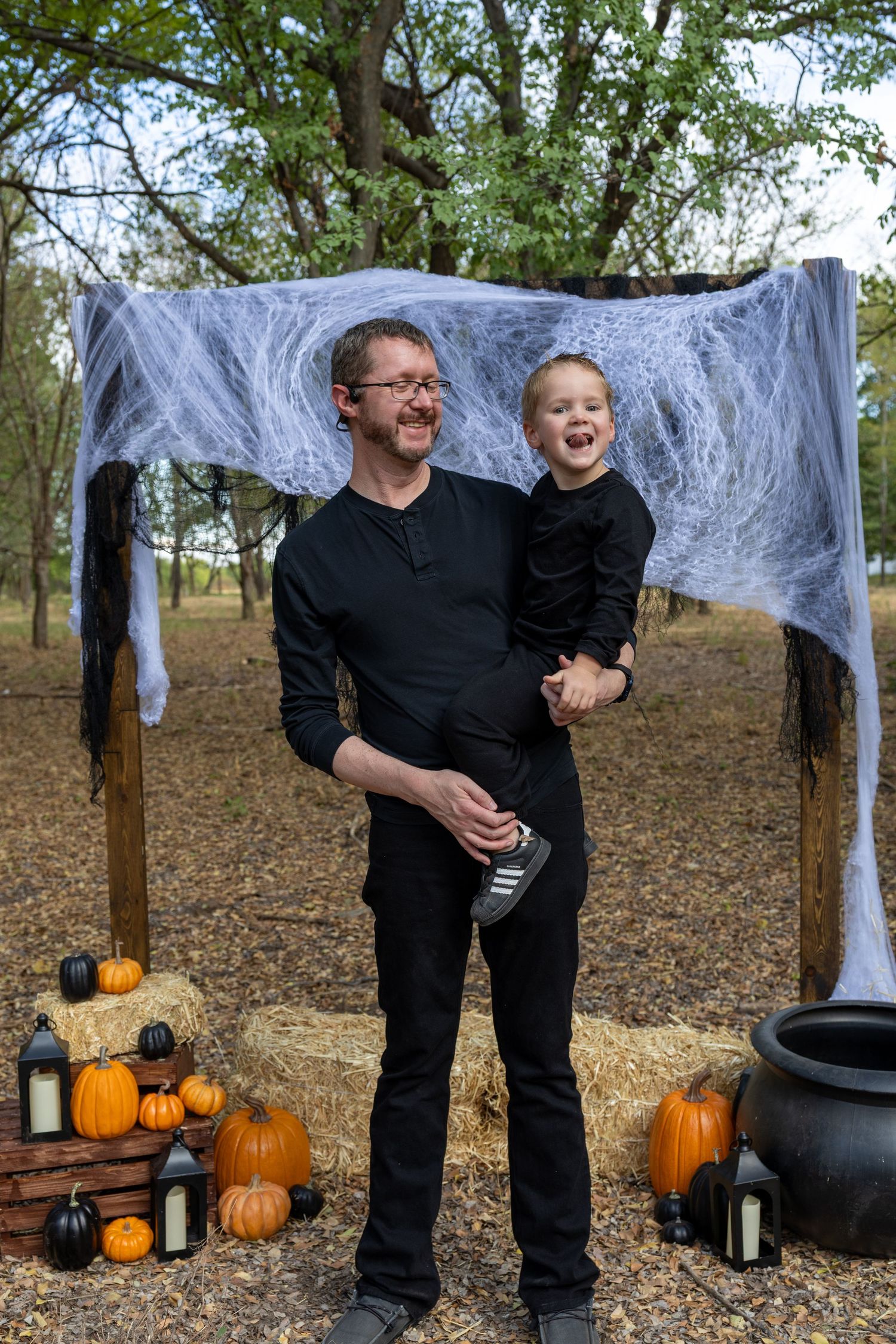 A man in black holds a laughing child at a Halloween outdoor display with pumpkins, hay bales, and spooky spider webs.