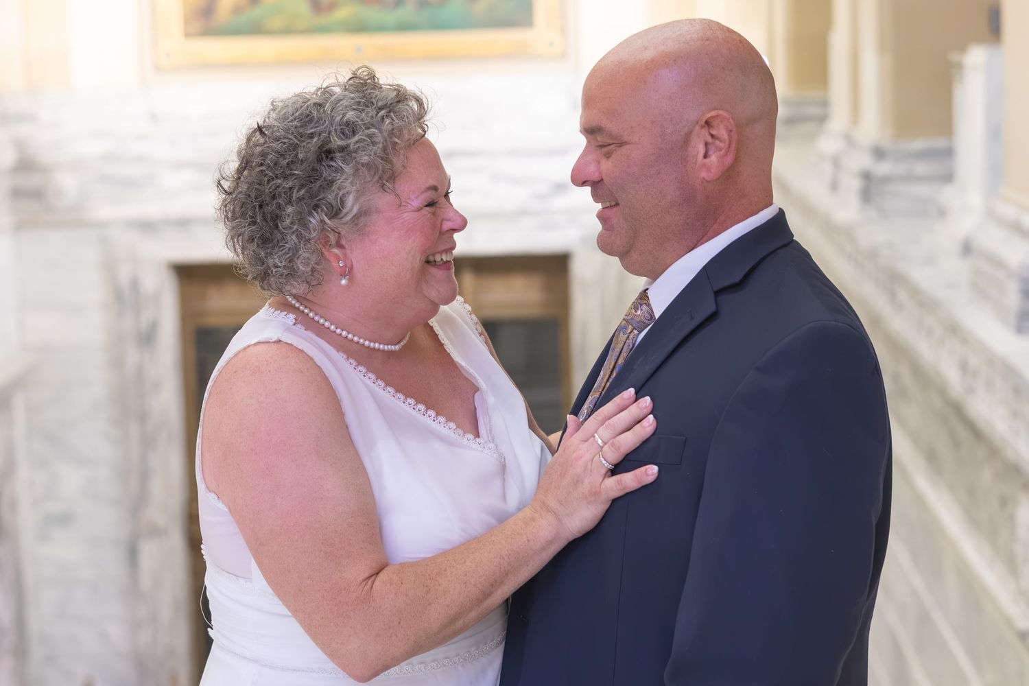 Smiling couple in wedding attire sharing a joyful moment, woman in white dress and man in navy suit, indoors.