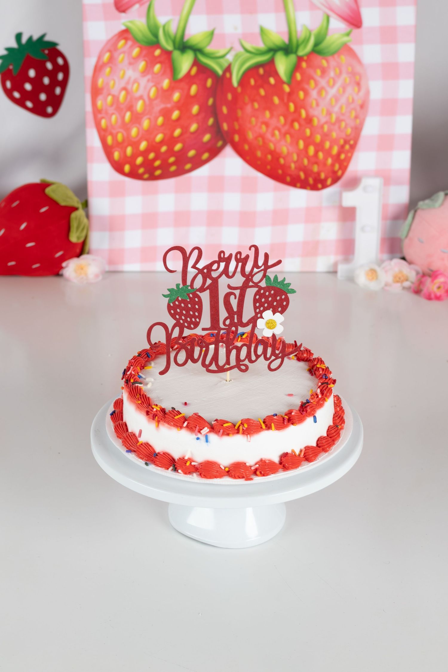 Strawberry-themed first birthday cake with Berry 1st Birthday topper on white cake stand with decorative backdrop.