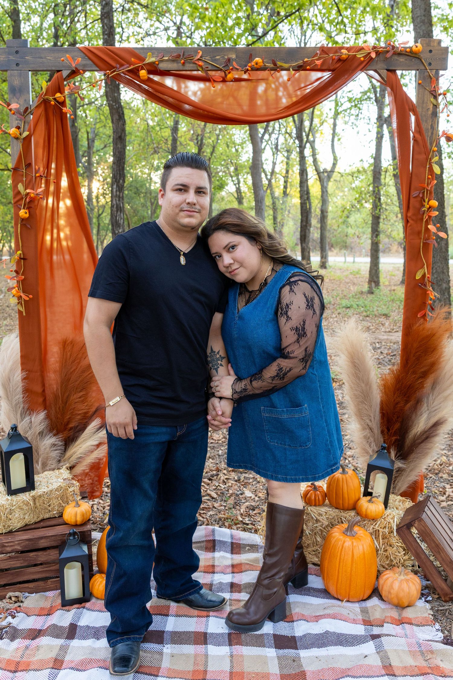 Couple posing together under a rustic fall arch with pumpkins, hay bales, and orange decor in an outdoor autumn setting.