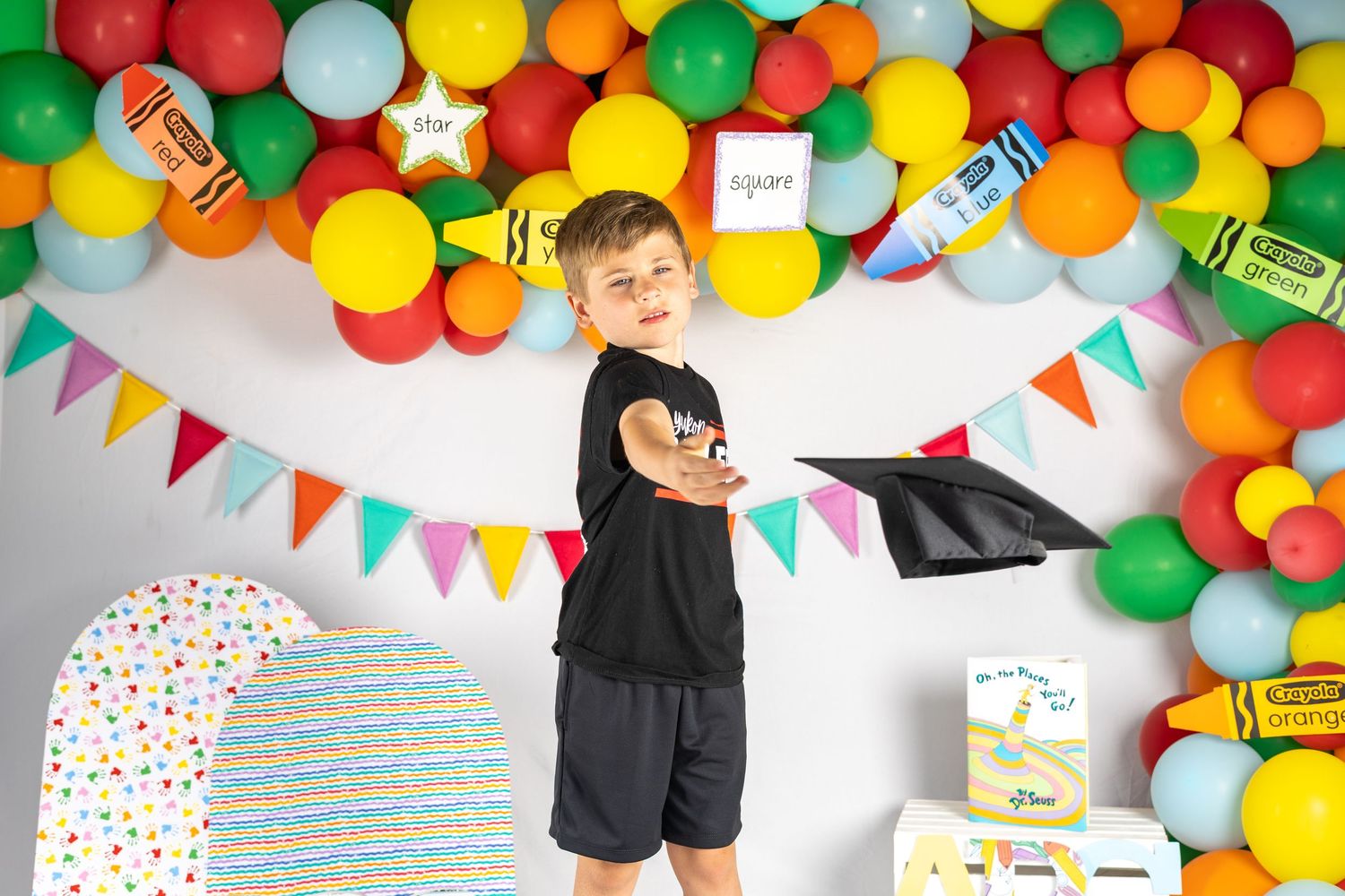 Boy in black outfit holding graduation cap, colorful balloon arch and bunting backdrop, crayon decorations.