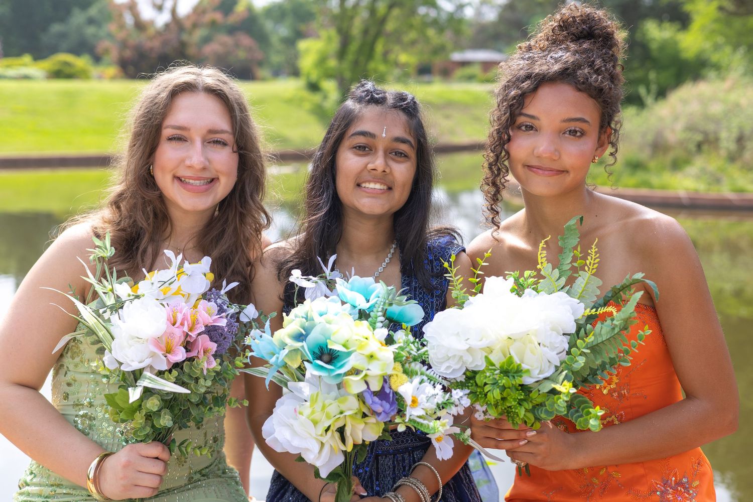 Three young women in formal dresses holding colorful flower bouquets outdoors near a pond on a sunny day.